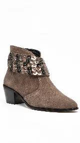 Bota corta de gamuza color visón, con taco medio ancho y punta afinada. Cuenta con un aplique desmontable alrededor del tobillo decorado con lentejuelas y apliques metalizados.