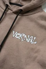 Hoodie color visón con calce relajado, capucha con cordones ajustables y bolsillo canguro frontal. Presenta un estampado pequeño en el pecho con el logo de la marca en estética hand-drawn.