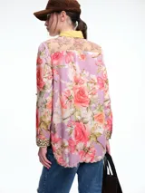 Camisa de silueta relajada con estampado floral en tonos rosas y violetas. Presenta cuello camisero en color amarillo mostaza, cierre frontal con botones, y puños con detalle de estampado animal print. El ruedo tiene un acabado festoneado.