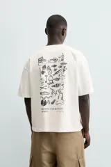 Camiseta de manga corta color blanco roto, corte relajado, con un pequeño bordado/estampado en el pecho que incluye una manzana, una rama de laurel, una trompeta y dos dados.