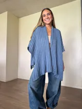 Ruana tejida de algodón con hilo lurex en punto jersey, color azul, con diseño amplio y detalle de etiqueta de cuerina en la parte posterior.