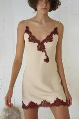 Vestido mini tipo lencero de tejido satinado color crema, con escote en pico y tirantes finos. Presenta detalles de encaje a contraste en color bordó en el escote y en el ruedo.