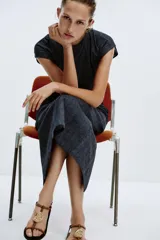 Sandalia plana con tiras de color marrón y un gran adorno metálico dorado circular en la parte frontal. Cuenta con cierre de hebilla ajustable al tobillo y suela plana de color negro.