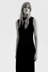 Vestido largo sin mangas confeccionado en velour suave. Presenta un diseño entallado con cuello alto y un efecto drapeado creado mediante pliegues estratégicos en el hombro y en el lateral de la cintura.