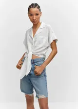 Camisa blanca de lino con cuello camisero, manga corta con vuelta, diseño crop y oversize. Tiene un bolsillo de parche en la parte delantera, cierre de botones y lazo ajustable.