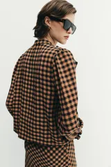 Blusa de manga larga con estampado de cuadros vichy en tonos marrón y negro. Presenta cuello redondo, cierre frontal con botones y detalle de volantes en el pecho.