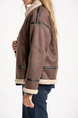 Campera tipo gamulán de corte oversize, color marrón con interior y cuello de corderito sintético en tono crema. Presenta detalles de ribetes en cuero sintético negro a lo largo de la prenda, cierre frontal metálico y puños tejidos.