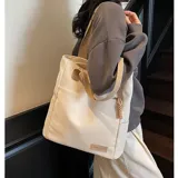 Cartera tipo tote de lona color marrón con asas de mano en color beige. Cuenta con un bolsillo frontal, bolsillos laterales y un detalle de tirador de cierre en cordón blanco.