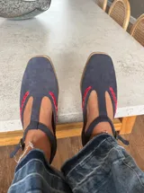 Ballerinas de gamuza color marrón oscuro con diseño tipo T-strap y detalles de costuras decorativas en color verde oliva. Cuentan con cierre de pulsera al tobillo y suela plana.