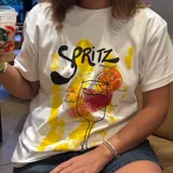 Remera blanca de corte clásico con estampa frontal ilustrada que representa un cóctel Spritz, incluyendo texto alusivo y detalles en tonos amarillos, naranjas y rojos con efecto de pinceladas.