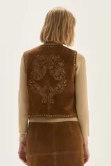 Chaleco de cuero color camel con bordados decorativos, tachas metálicas en los bordes y cierre frontal mediante lazos.