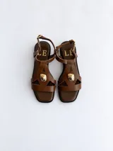 Sandalias planas de cuero color marrón, con punta cuadrada, tiras con recortes geométricos y aplique metálico dorado en la parte frontal. Cuenta con tira ajustable al tobillo con hebilla dorada.