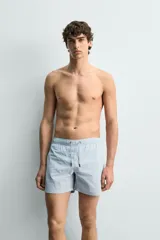 Bañador de hombre de largo medio, color celeste claro con rayas blancas horizontales. Presenta cintura elástica ajustable con cordón, bolsillos frontales y bolsillos traseros de plastrón. Incluye forro interior.