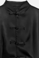 Chaqueta negra de tejido satinado, con cuello mao, manga larga con hombreras, corte evasé y cierre frontal con alamares.