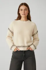 Sweater de tejido de punto grueso color gris oscuro, con cuello redondo a la base y mangas largas. Presenta detalles de bordado decorativo en color blanco con diseño de cruces en las mangas y el ruedo.