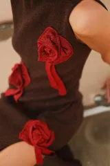 Vestido midi marrón de lurex, con cuello redondo y sin mangas. Presenta apliques de rosettes rojas en el frente y tajo lateral.