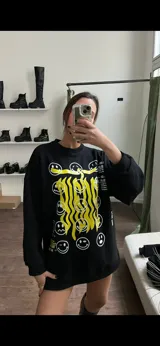 Buzo negro de corte amplio con cuello redondo y mangas largas. Estampado frontal con caritas sonrientes y texto en amarillo.