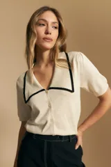 Top de tejido color beige claro, manga corta, escote en V con cierre frontal de botones y ribetes negros en el cuello y el borde del escote.