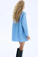 Vestido camisero de corte evasé y holgado, color celeste con finas rayas verticales en negro. Presenta cuello de pico, botonadura frontal y mangas largas con puños abotonados.