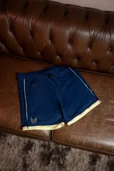 Short deportivo de lycra color azul marino con cordón de ajuste negro. Presenta ribetes y un pequeño logo de alas en color dorado/beige en la parte inferior izquierda.