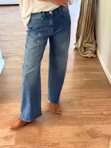 Pantalón de jean azul con corte holgado y detalle de tres cruces bordadas en el muslo derecho. Presenta costuras reforzadas y cierre frontal con botón metálico.