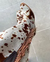Botas texanas de caña media confeccionadas en cuero vacuno con estampado animal print tipo vaca. Presentan flecos laterales en gamuza marrón, taco cuadrado bajo y puntera ligeramente redondeada.