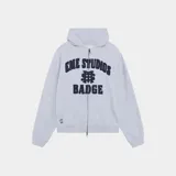 Campera con capucha de felpa gris melange, con cierre frontal de doble cremallera metálica, bolsillos tipo canguro y estampado frontal con el texto "Eme Studios Badge" en azul marino.