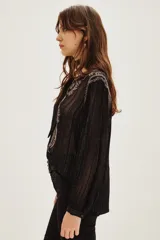 Camisa de manga larga en color negro con tejido semitransparente y detalles de hilos metalizados. Presenta un diseño con bordados florales en tonos tierra, cierre frontal con botones y un lazo decorativo en el cuello.