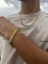 Conjunto de joyería de color dorado, compuesto por una cadena de eslabones tipo cubana o espiga y una pulsera a juego con eslabones anchos y planos. El conjunto se luce sobre una camiseta beige de manga corta con texto estampado en negro.