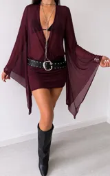 Vestido corto de tul color bordó con escote profundo en V y mangas largas acampanadas con puños ajustados. Presenta un diseño traslúcido y fluido.