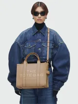 Cartera tipo tote de cuero granulado color tostado, con diseño estructurado y rectangular. Presenta doble asa superior fija y una correa larga ajustable y extraíble. En el frente, destaca el logo estampado con la inscripción 'THE TOTE BAG MARC JACOBS'.