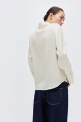 Blusa blanca de tela con brillo, cuello clásico, escote en V con botones frontales, mangas largas con puños sencillos y bajo redondeado.