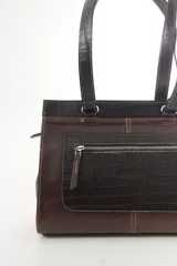 Cartera de cuero marrón de tamaño medio con diseño estructurado, que combina cuero liso y un panel frontal texturizado tipo cocodrilo con bolsillo exterior con cierre. Cuenta con doble asa de hombro y costuras visibles de contraste.