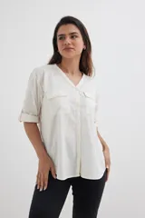 Camisa color blanco roto, de manga corta enrollada a 3/4, con escote en V y cierre frontal de botones. Presenta dos bolsillos de parche con solapa en el pecho.