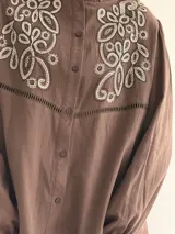 Camisa color marrón chocolate con bordado artesanal de flores y hojas en hilo crudo. Presenta cuello con volado, mangas largas con puños abotonados y cierre frontal con botones.