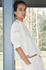 Blusa de algodón color blanco con cuello redondo y mangas abullonadas. Presenta detalles de puntilla a tono en el pecho y las mangas, bajo recto y cierre posterior con abertura y botón.