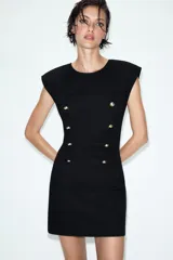Vestido corto de estilo militar en color negro, con cuello redondo, hombreras y manga sisa. Presenta un diseño frontal con falsos botones dorados dispuestos en filas horizontales. Cierre lateral con cremallera oculta y abertura en la espalda con botón.