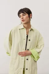 Chaqueta oversize de gabardina color verde claro, con solapa clásica, bolsillos frontales, cierre de botones oscuros y mangas largas con vuelta en tono verde lima vibrante.