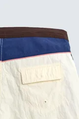 Short de baño blanco marfil con cintura marrón y cordón ajustable del mismo color. Presenta franjas laterales en tono azul marino con finos bordes rojizos y bolsillos laterales.