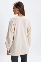 Buzo deportivo de corte oversize en color crudo, con cuello redondo y mangas largas. Presenta un diseño bordado en el frente con la inscripción Saint Tropez y un motivo floral central.