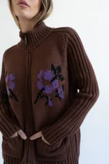 Cardigan de punto color marrón con cierre frontal, cuello alto y bolsillos aplicados. Presenta diseño tejido localizado con detalles en violeta y negro en el pecho.