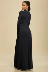Vestido largo de manga larga en color negro, confeccionado en tela elastizada. Presenta un diseño con escote profundo y detalle de abertura superior, con drapeado central en la cintura que estiliza la silueta y una abertura frontal en la falda.