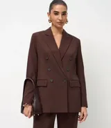 Blazer de sastrería color marrón, de corte regular y largo, con cuello peaked, cierre frontal cruzado con botones, bolsillos con tapa y mangas largas con botones decorativos.