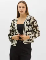 Cardigan abierto de tejido crochet con diseño de flores bicolores en negro y crema. Presenta mangas largas, frente abierto sin cierre y bolsillos frontales tipo parche. Posee terminaciones con franjas a contraste en escote, puños y ruedo.