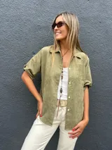 Camisa de gamuza sintética liviana en color verde oliva, con cuello clásico, cierre frontal de botones y mangas cortas con dobladillo.