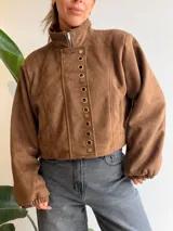 Chaqueta corta de efecto gamuza con cierre frontal y botones. Presenta mangas tipo globo con puños elásticos y detalle de aros metálicos decorativos en la solapa. Cuenta con ajuste lateral en la cintura y forro interior.