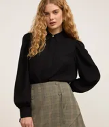 Camisa de crepé color negro, con cuello inglés, cierre frontal abotonado y mangas largas abullonadas con puños ajustados.
