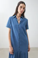 Maxi vestido camisero de jean azul con cuello clásico, manga corta, cierre frontal con botones y falda amplia con volantes.