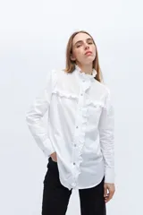 Camisa blanca de algodón con cuello alto con volados y bordado calado en el frente. Tiene mangas largas con puños abotonados y cierre frontal con botones.