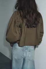 Camisa de popelín color verde militar, con cuello solapa, mangas largas abullonadas terminadas en puño, bajo recto y cierre frontal con botones ocultos por solapa.